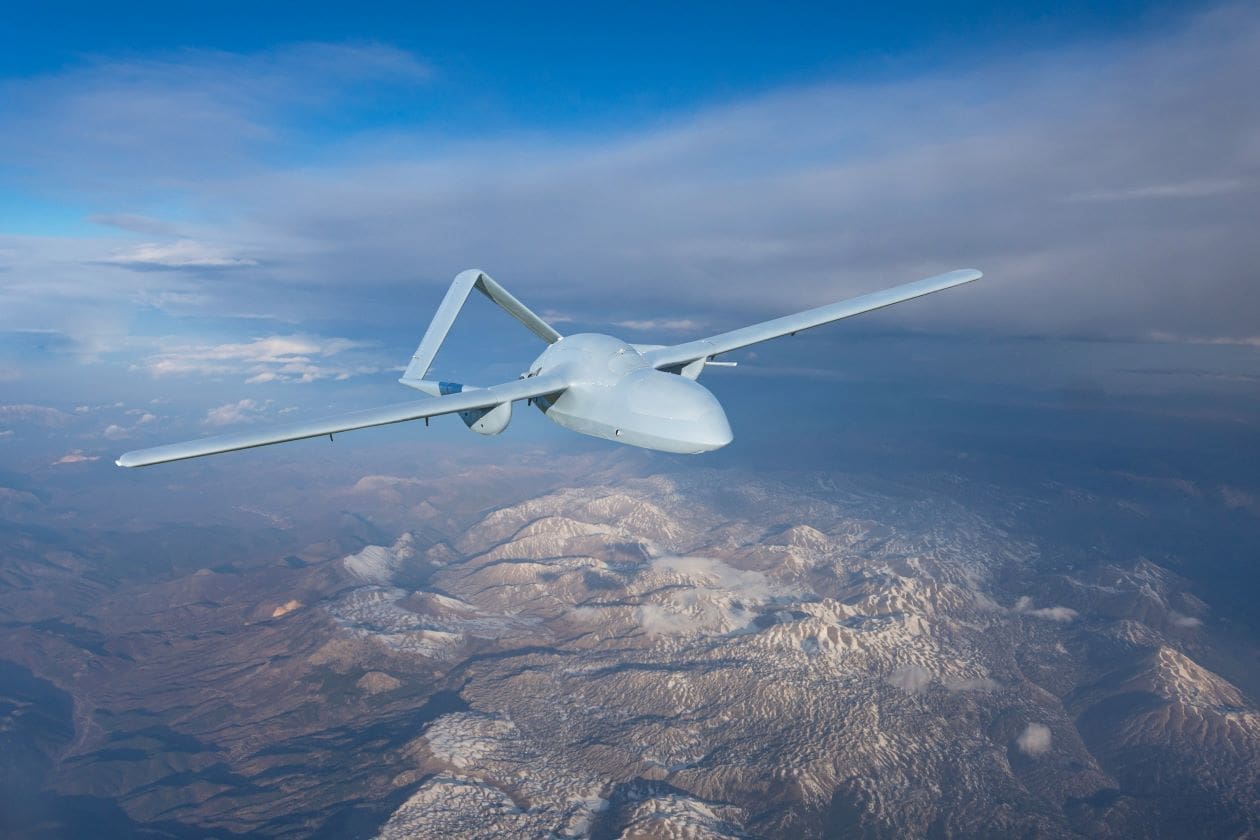 A white drone with wide wings flies over a mountainous landscape with snow-covered peaks under a cloudy sky.