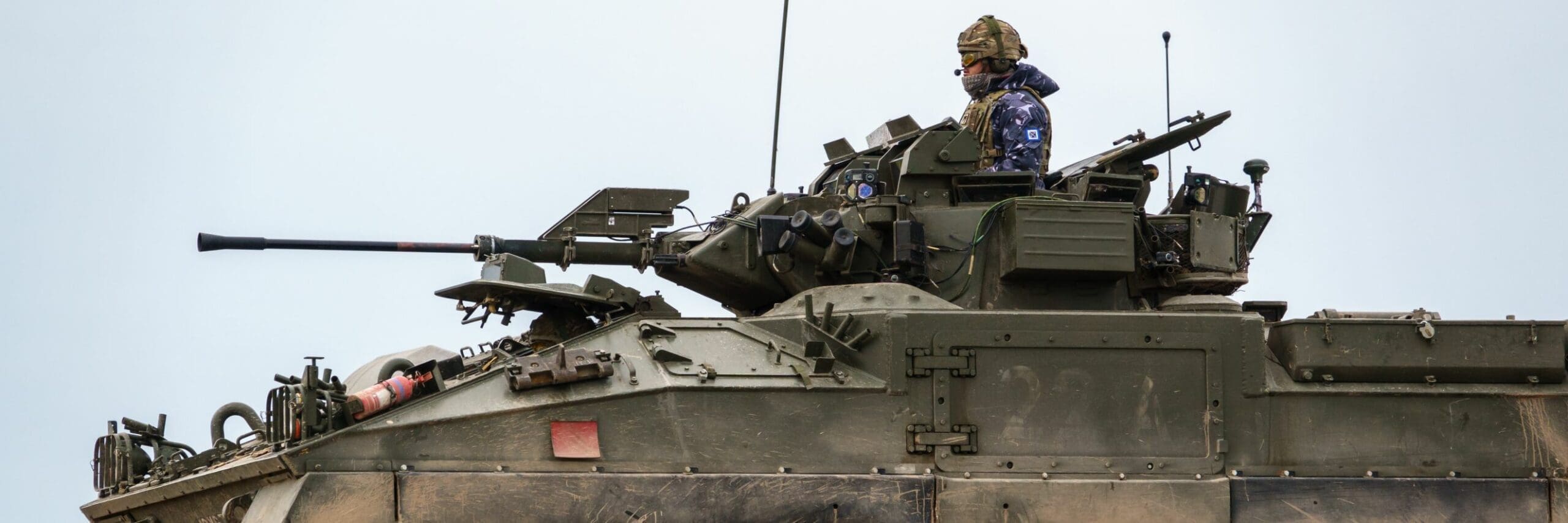 Soldier in camouflaged uniform standing atop an armored military vehicle with mounted weaponry, ready to face the latest trends in warfare.