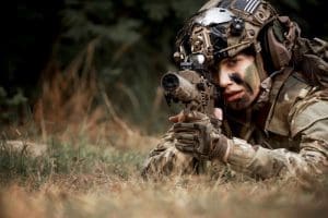 A soldier in camouflage and tactical gear, lying on the ground and aiming a rifle with a serious expression, epitomizes the latest trends in the defense industry’s advances in security.