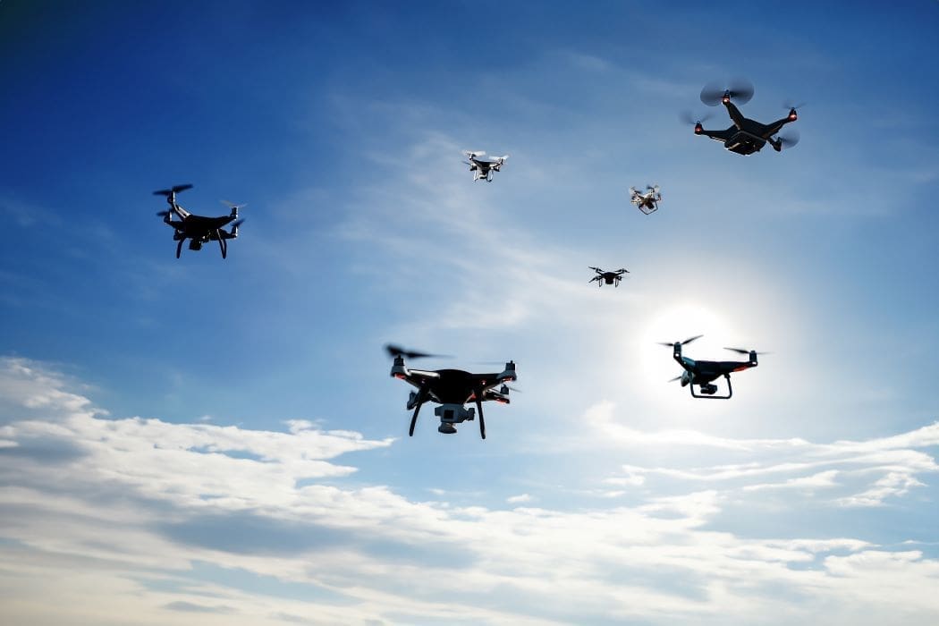 A swarm of drones flies in the sky against a backdrop of clouds and sunlight, hinting at the future of warfare.