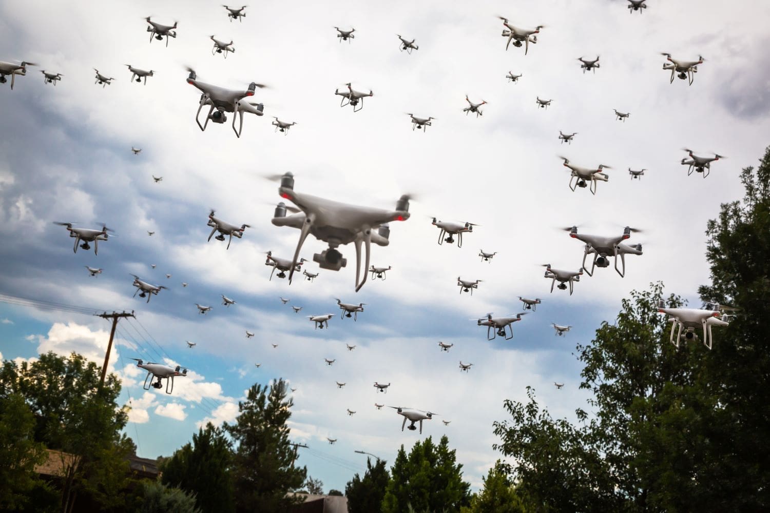 A large group of drones flying in the sky over a tree-lined area with a power line visible in the background. The sky is cloudy.